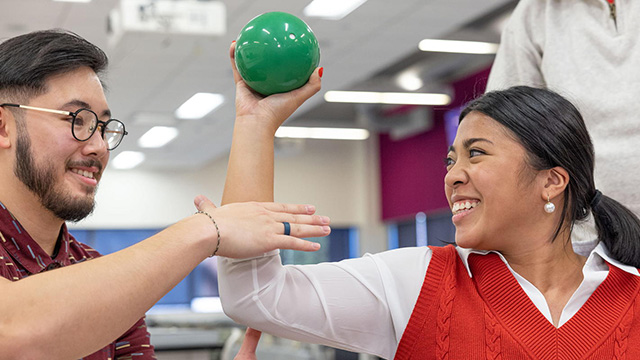 Two students performing a physical therapy exercise, one is holding a green ball in their hand