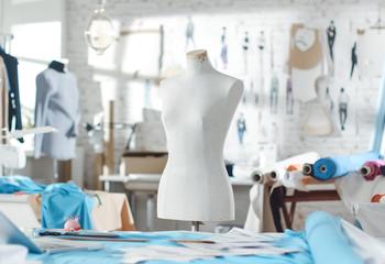 Shot of a Tailoring Mannequin that Stands in a Bright and Sunny Studio. Various Sewing Items and Colorful Fabrics Laying around, Mannequins Standing, and Sketches Pinned to the Wall.