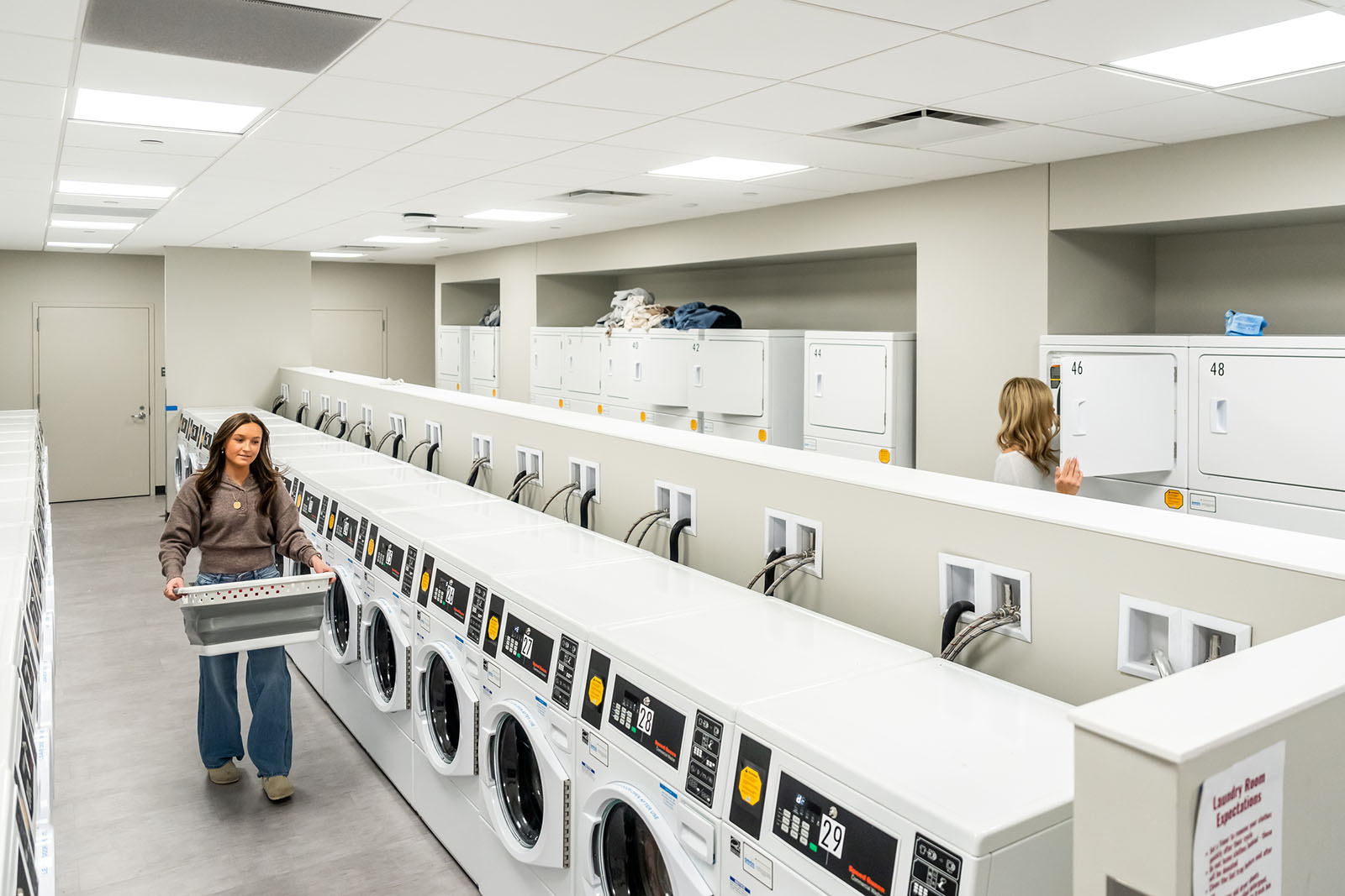 A student holding a laundry basket walking past a row of washing machines