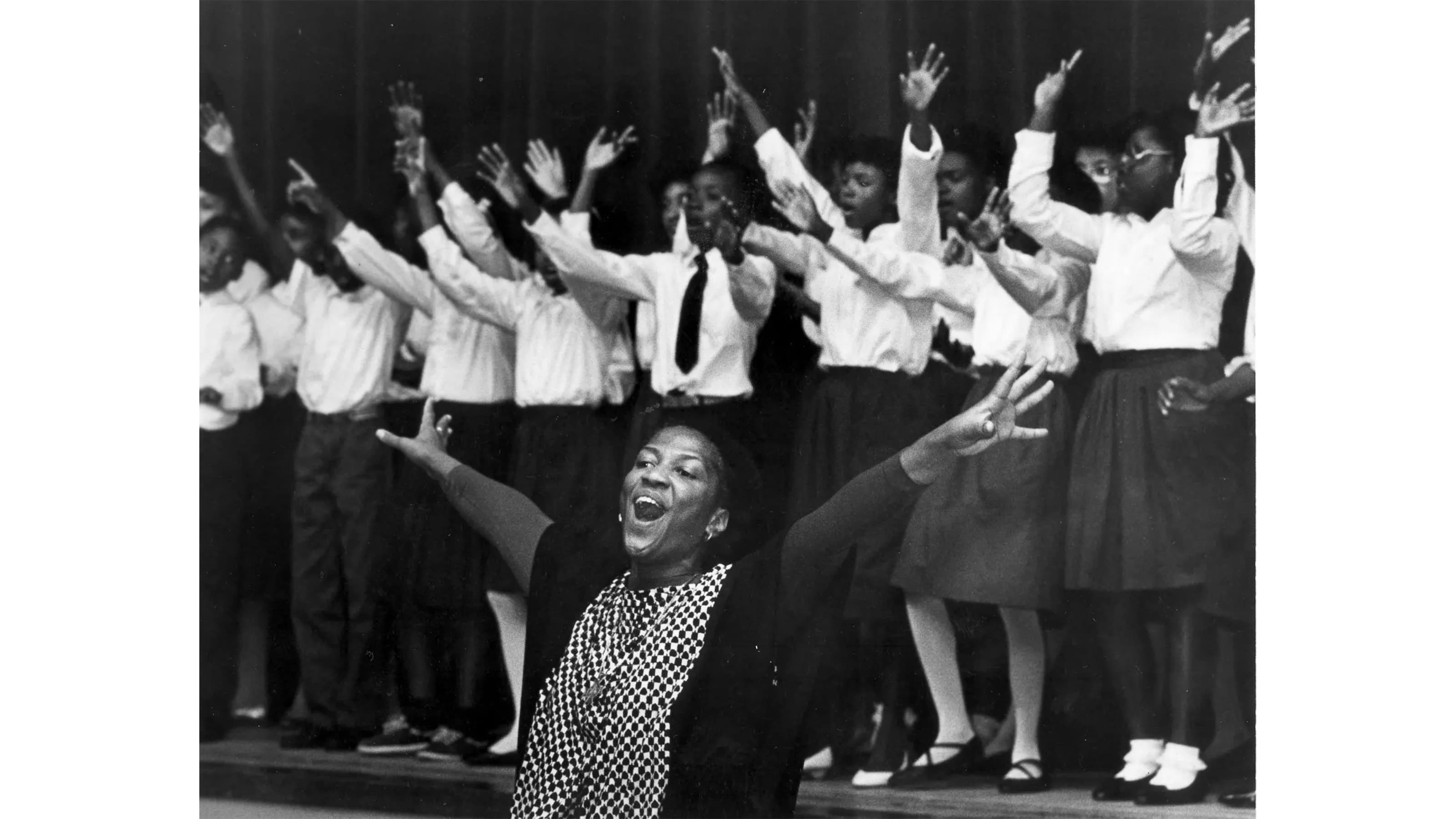 A woman with her arms outstretching singing with a chorus of people behind her