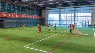 An indoor field with players dressed in crimson uniforms playing lacrosse