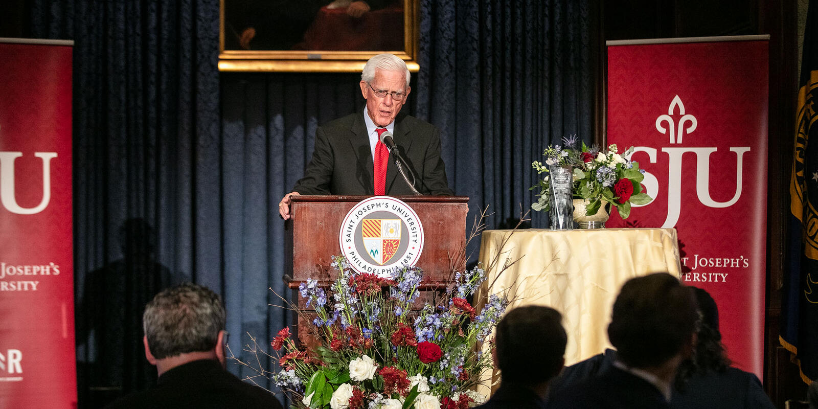 A man standing at a podium speaking to an audience with red banners behind them