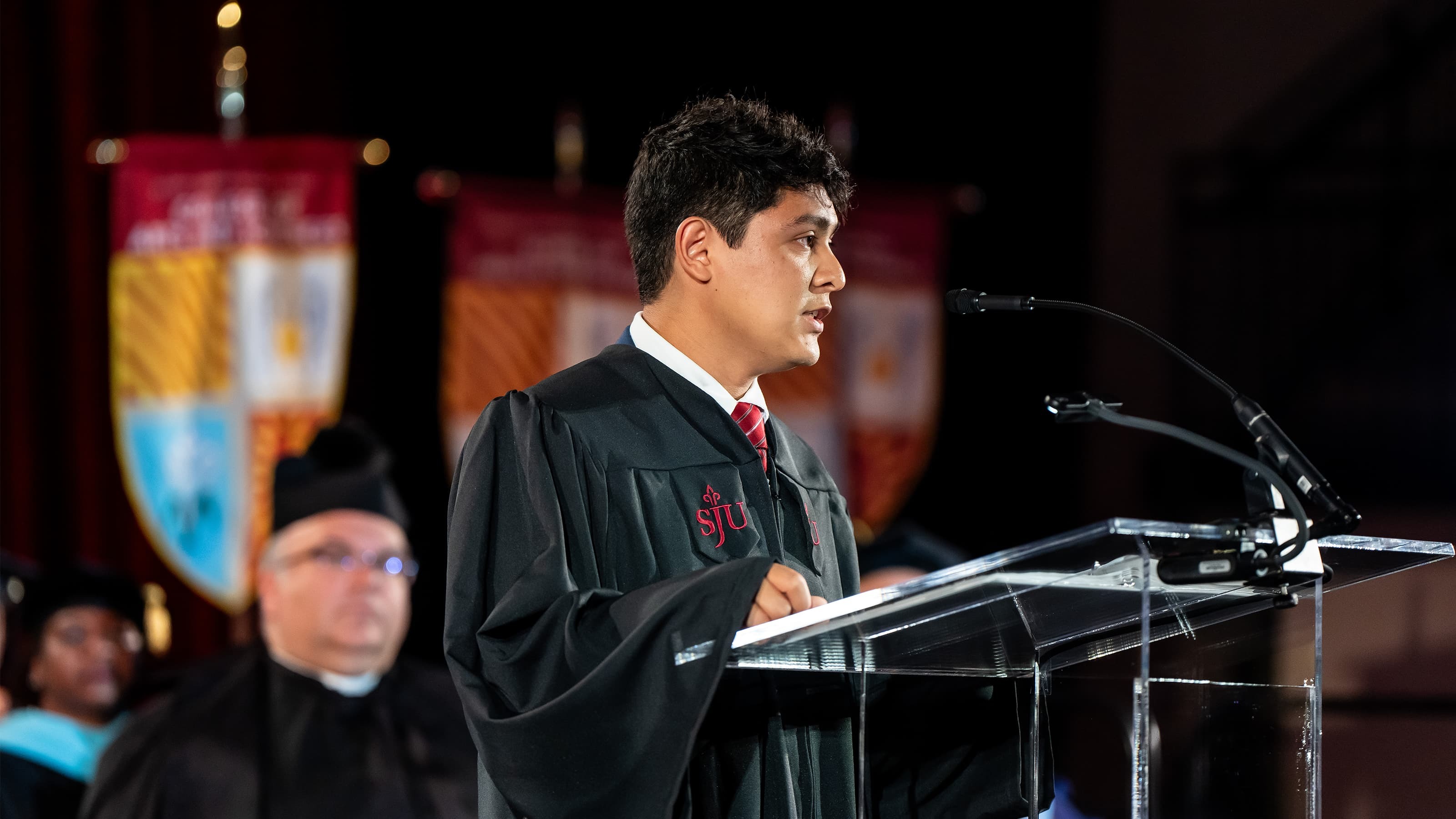 Milton O'Brien, BS ’25, speaks into a microphone in a black robe. 