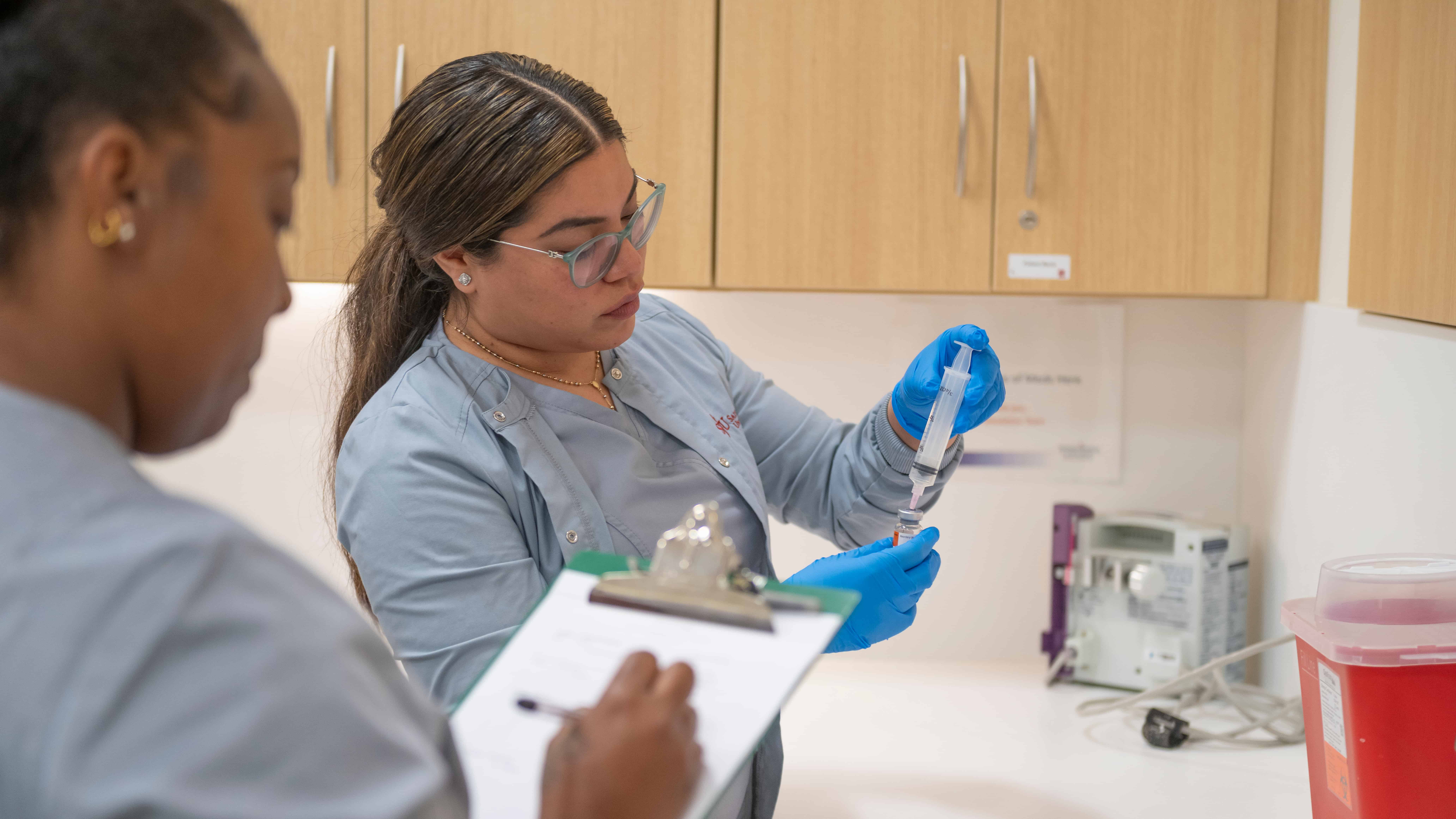 One nursing student holds a syringe while the other watches while writing on a clipboard.