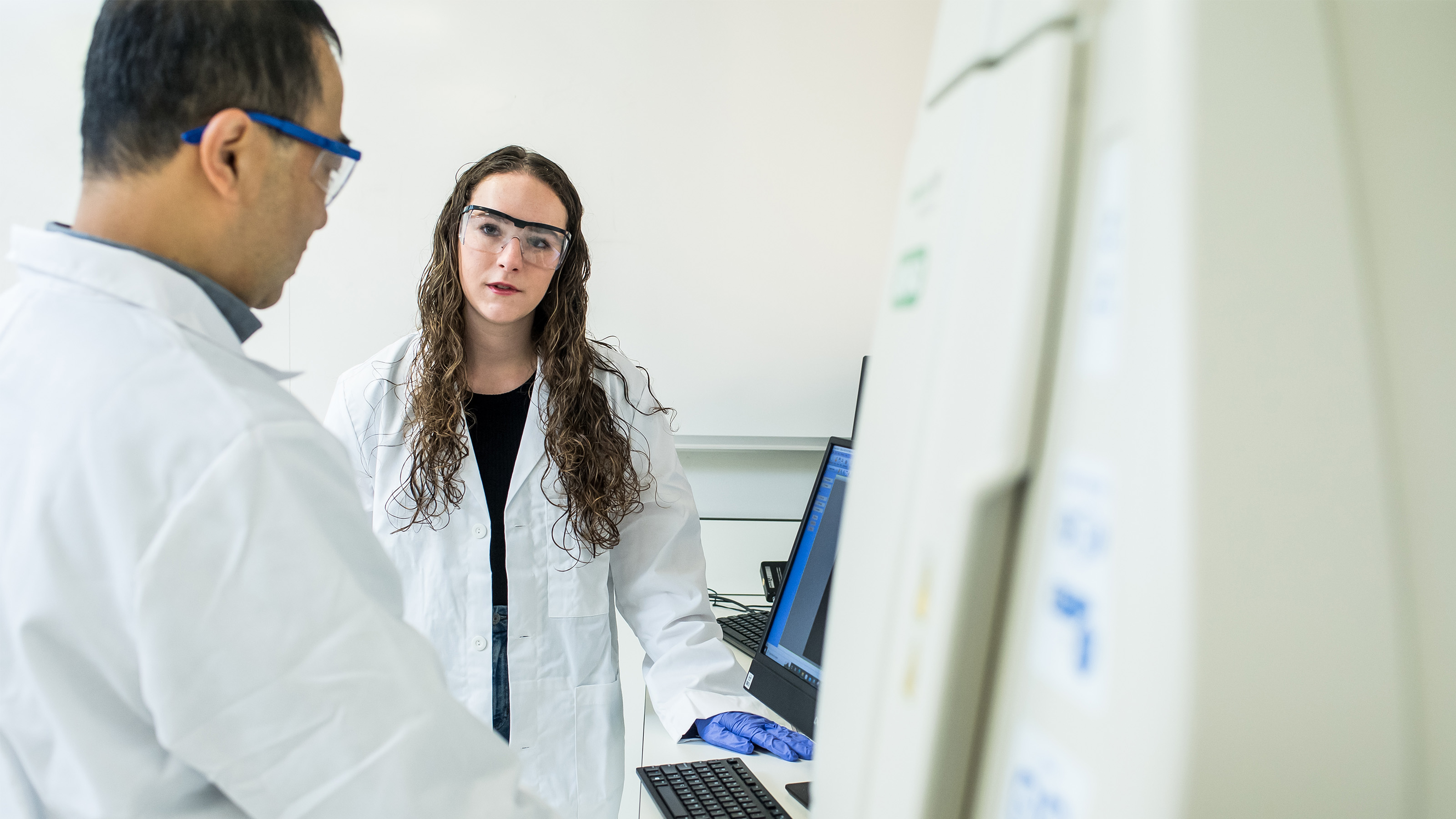 Student and faculty member wearing lab coats and safety goggles standing next to a computer