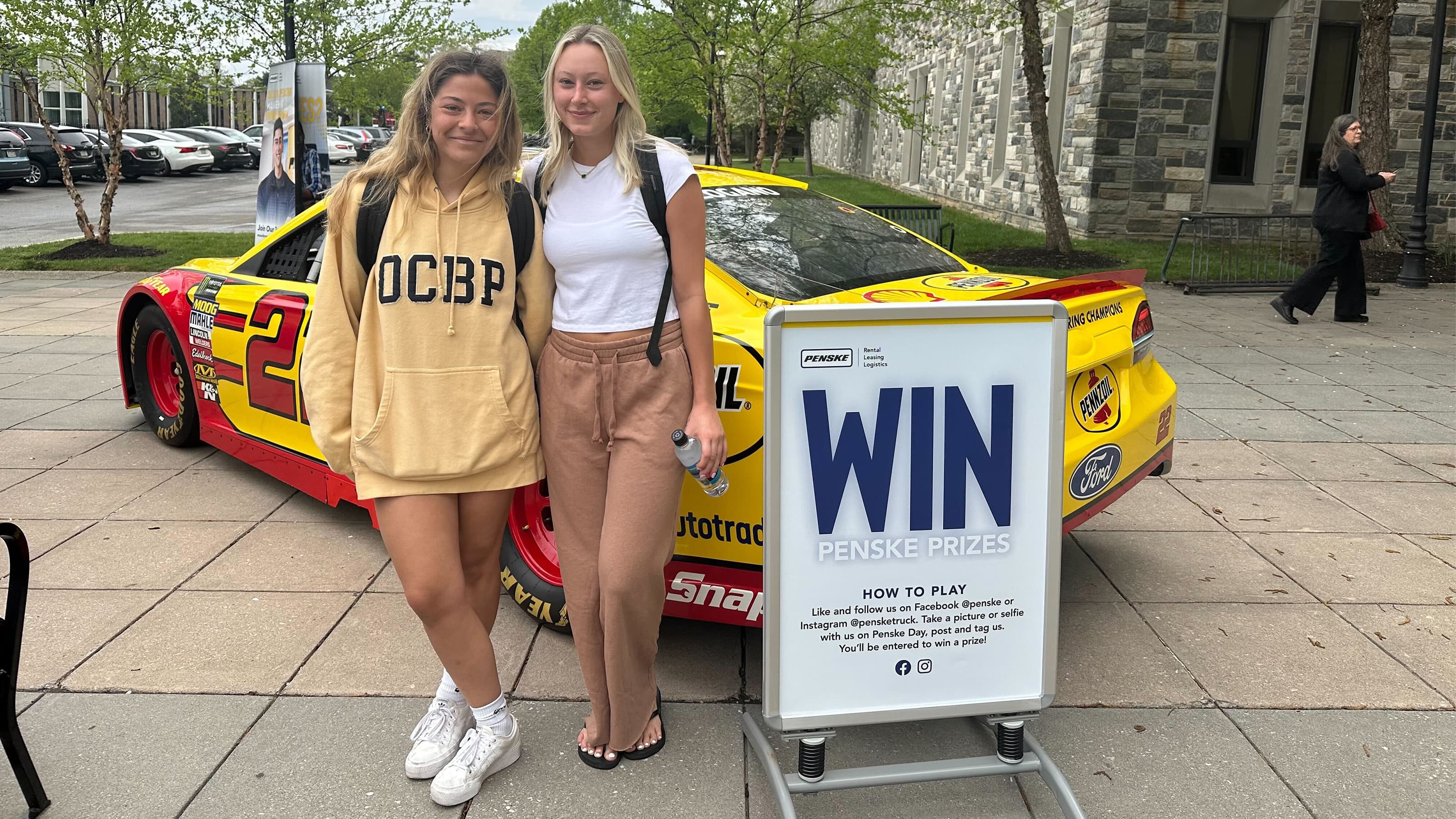 Two female students standing in front of a Penske racecar