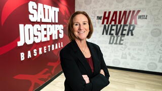 Jill Bodensteiner standing in front of red Saint Joseph's basketball wall on her left side and white 'the Hawk Will Never Die' slogan on her right