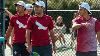 Three male athletes wearing red shirts outside on a tennis court