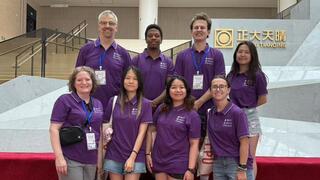 Students and faculty members in purple shirts smile in front of a silver display.