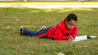 An elementary school student in a red hoodie completes his classwork on the grass.