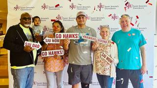 A family standing in front of a Saint Joseph's University backdrop with signs that say "SJU Mom," "SJU Dad," "Go Hawks" and "#HawkHillBound"