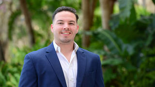 Headshot of Jake DeMarco in blue suit standing outside in front of trees