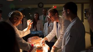 Male professor and two students in lab coats working with high-fidelity mannikin with students observing in the background