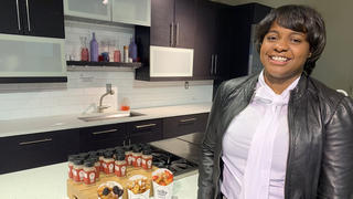 Charisse McGill stands in a white kitchen. On the counter is a board filled with bottle of French toast spice.