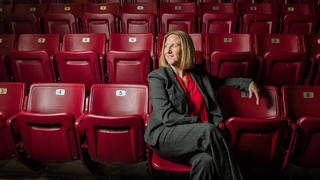 Jill Bodensteiner, in a gray suit, sits among a sea of empty red seats. She looks off-camera.