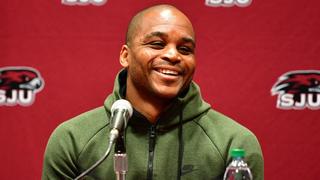Jameer Nelson wears a green track suit and laughs behind a microphone at a press conference.