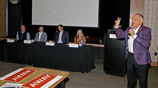 At left, three experts sit at a table. At right, Michael Solomon stands with a microphone, speaking to an unseen audience.