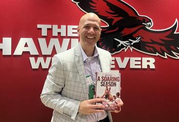 Aaron Bracy, BS ’98, MS ’99, poses with his book in front of the Hawk logo.