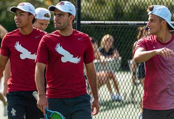 Three male athletes wearing red shirts outside on a tennis court