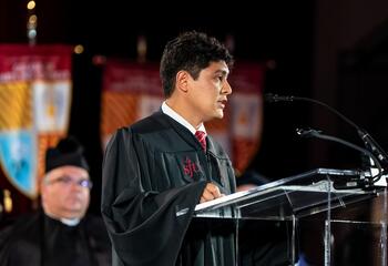 Milton O'Brien, BS ’25, speaks into a microphone in a black robe. 