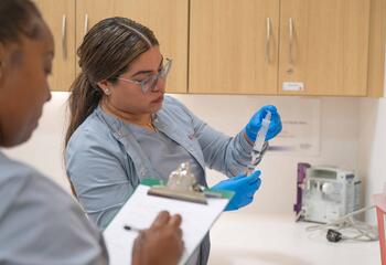 One nursing student holds a syringe while the other watches while writing on a clipboard.