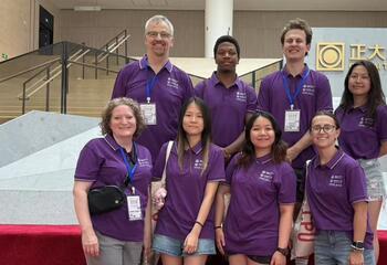 Students and faculty members in purple shirts smile in front of a silver display.