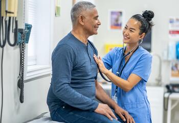 A nurse in blue scrubs listens to a patient's heart with a stethoscope.