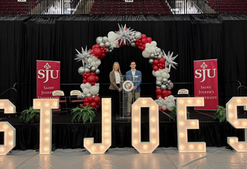 Stefano Billis, BS ’26, and Cheryl A. McConnell, PhD, on stage under a balloon arch with light up St. Joe's letter's in front of them.