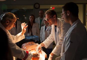 Male professor and two students in lab coats working with high-fidelity mannikin with students observing in the background
