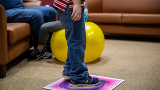 Little boy, wearing jeans, standing on a pink square with a yellow ball in the background.