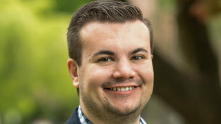 Will Marsh smiles at the camera wearing a suit jacket with trees behind him