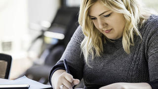 A woman in a grey sweater and glasses writes in a notebook