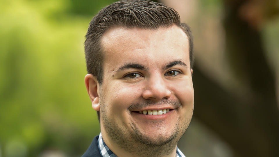 Will Marsh smiles at the camera wearing a suit jacket with trees behind him