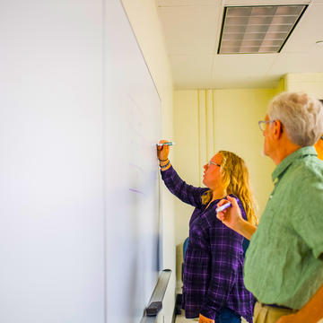 A woman writes on a white board with her professor