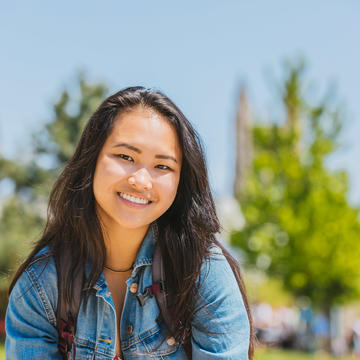 A woman smiles at the camera in a jean jacket with the campus behind her