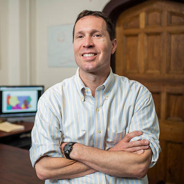 Kristopher Tapp, mathematics professor, smiling in an office with his arms crossed