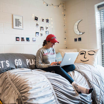 student in her dorm room, sitting on a bed with a laptop on her lap