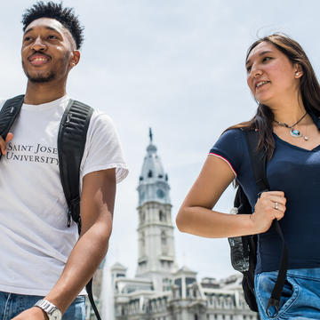 A male and female student are walking in the city, city hall is behind them in the distance