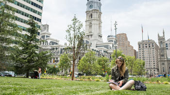 A woman sits on the grass looking at a book with part of the city skyline behind her.