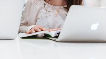 A close-up of a student's hands laying on a book next to a laptop