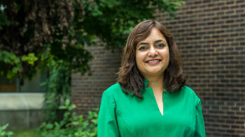 Usha Rao smiles wearing a green top in front of foliage and a brick wall