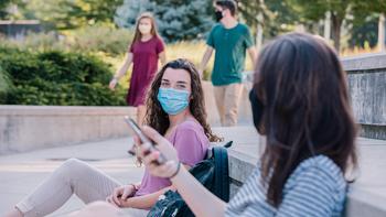 Two students sitting on a bench chatting, wearing face masks and holding phones, while two students walk in the background.