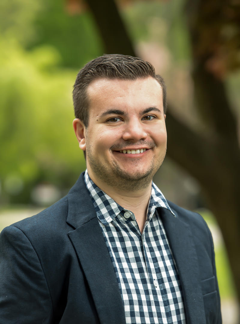 Will Marsh smiles at the camera wearing a suit jacket with trees behind him
