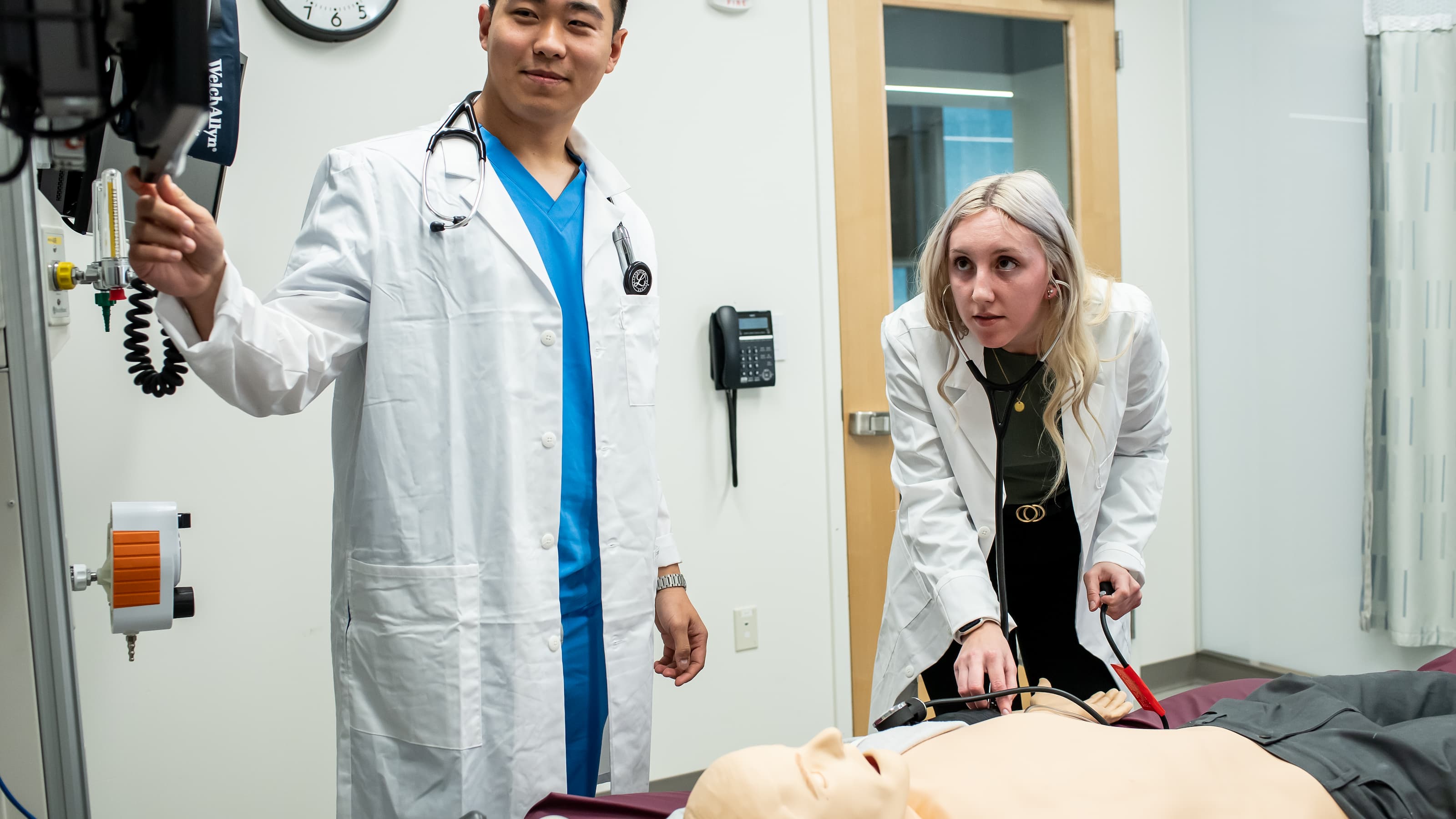 Two students in white lab coats practice a medical exam.