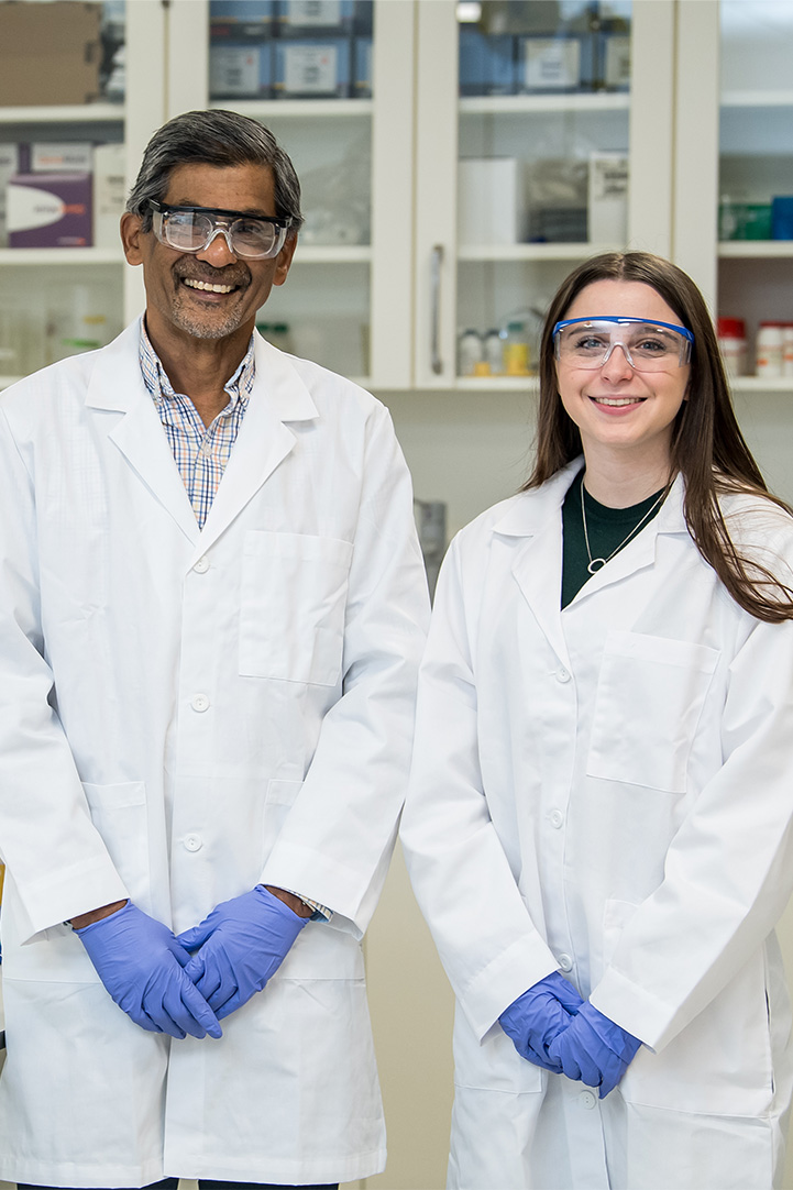 Student and faculty member wearing a lab coat and safety goggles pose for a photo in the lab