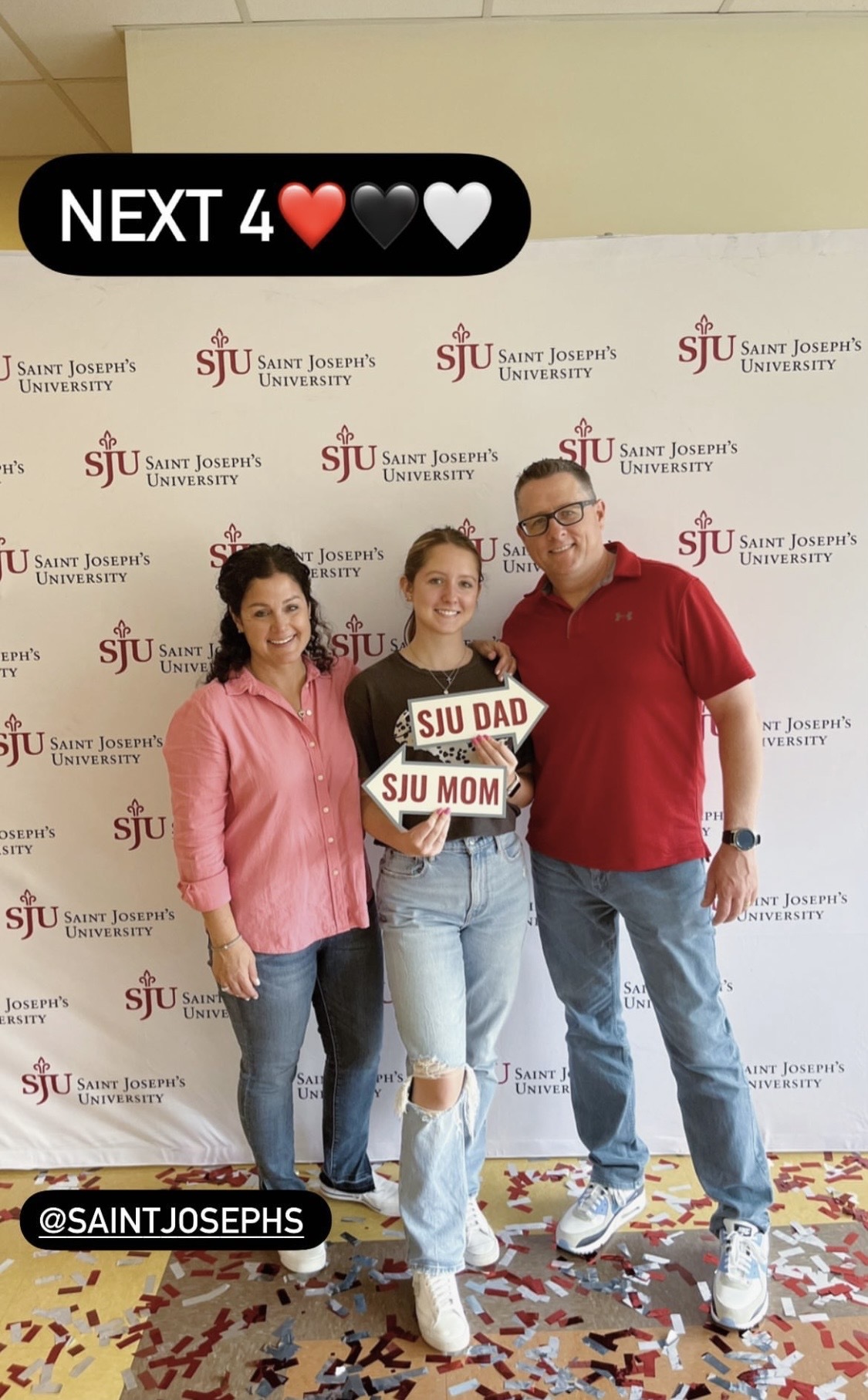 Student standing with her parents in front of a Saint Joseph's University backdrop with signs that say "SJU Mom" and "SJU Dad"