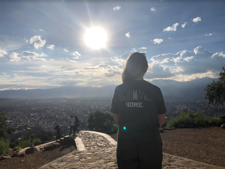 A person standing with the back to the camera wearing a shirt of a city skyline and the word "Home" across the middle. The person stands in front of a landscape of a mountain scape and city in South America.