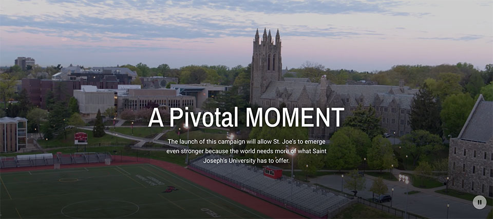 An aerial photo of Saint Joseph's University Hawk Hill campus complete with green athletic fields, large academic buildings, and gothic Barbelin Hall with the phrase A Pivotal Moment emblazoned in the foreground