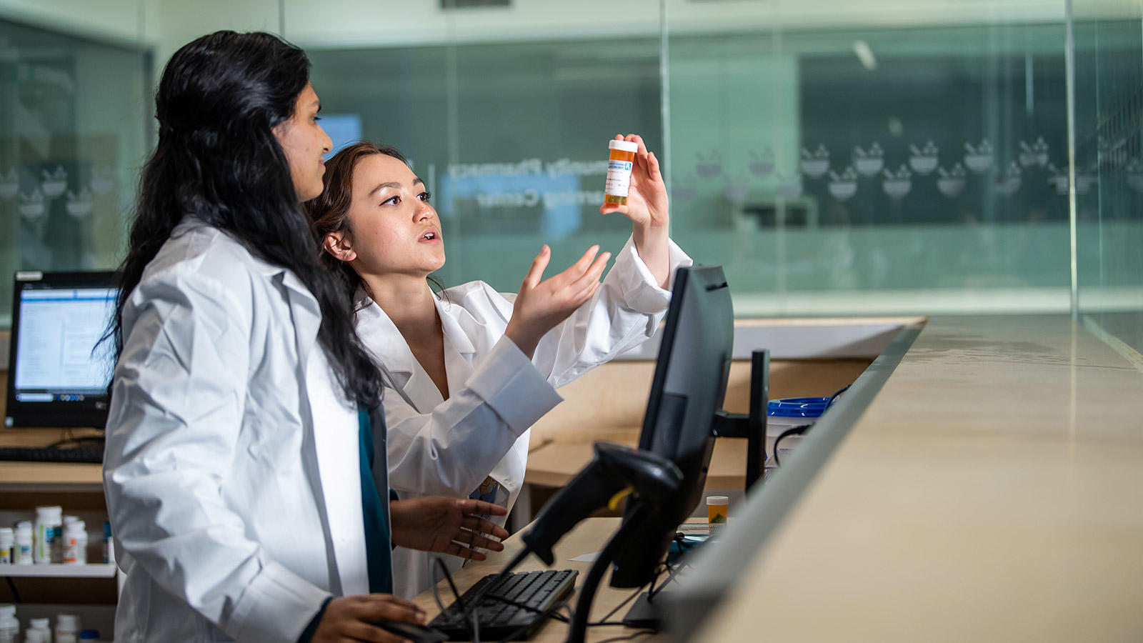 Students in white coats holding up a pill bottle in a pharmacy