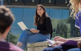 A smiling student sits outside Post Learning Commons with her laptop